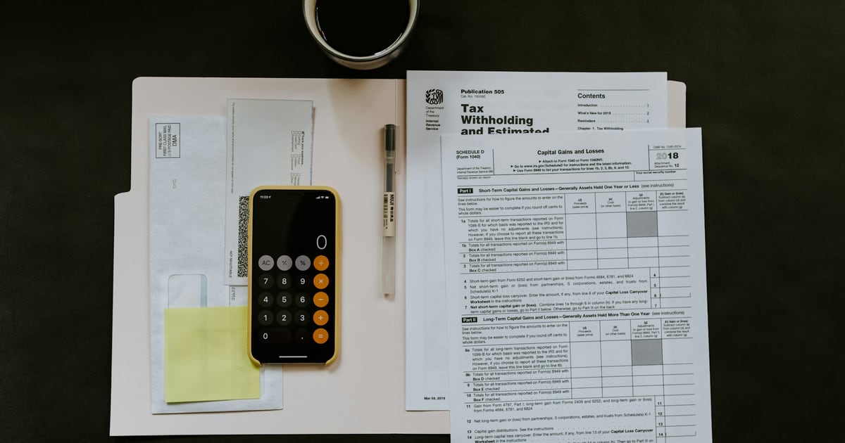 US tax withholding and estimated tax paperwork spread on a dark table beside a phone showing a calculator and a pen