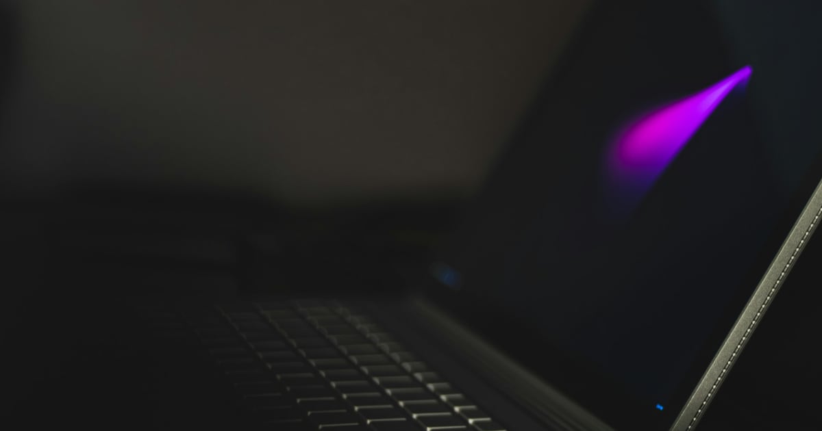 A laptop in near darkness, its screen dark and turned away, with only a faint violet glare on the lid