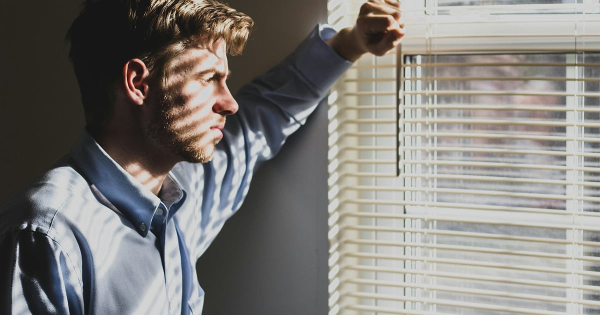 A man leaning on a window frame in low light, stripes of light from the blinds across his face, looking away
