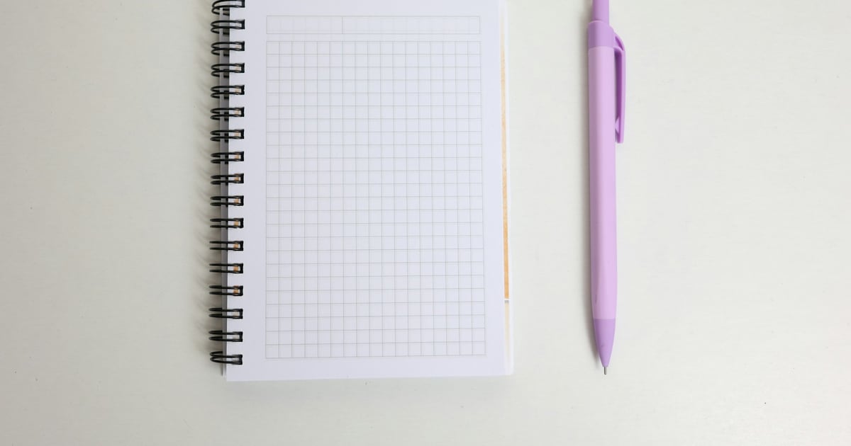 A blank spiral-bound grid notepad next to a lilac pen on a plain white surface