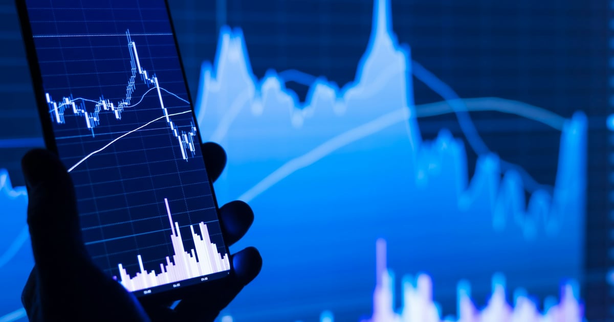 A hand holding a phone with a stock chart on it in front of a large blue chart on a screen behind