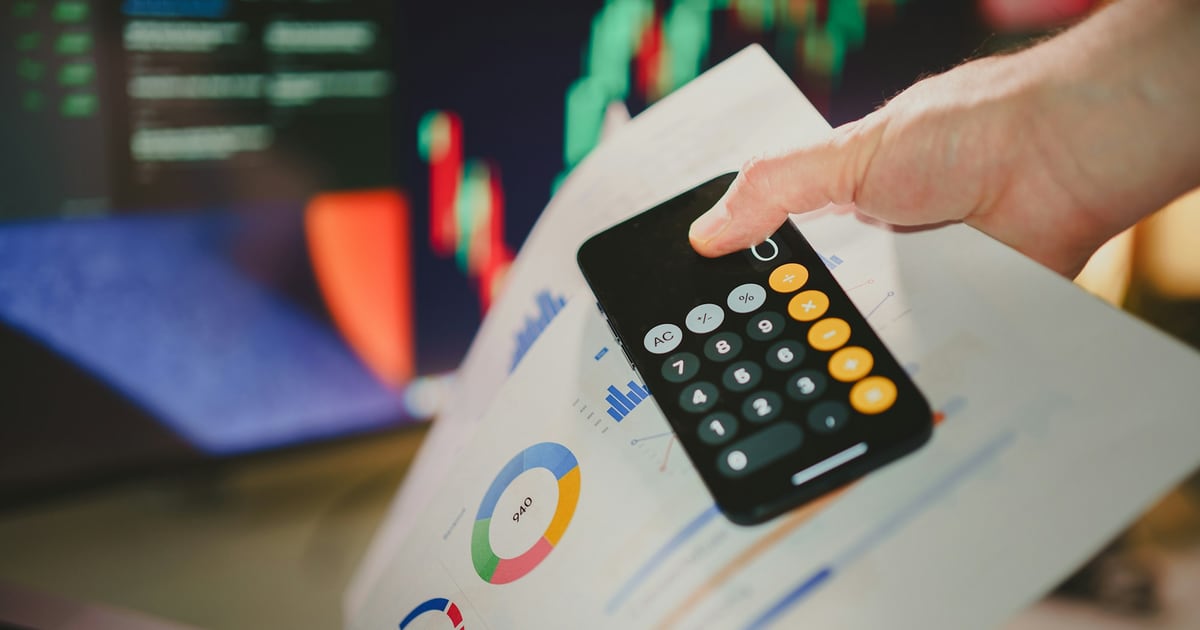 A hand holding a phone calculator over printed charts, with a colored trading screen blurred in the background