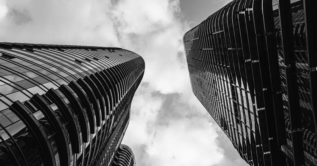 Black and white view looking straight up between two ribbed skyscrapers into a cloudy sky
