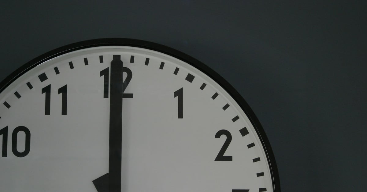 Close crop of a large wall clock face with both hands pointing straight up at twelve
