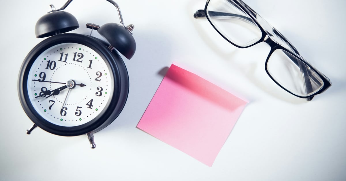 A black twin-bell alarm clock beside reading glasses and a pink sticky note on a white desk