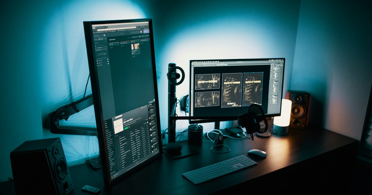 A desk lit in teal with one vertical monitor and one horizontal monitor, a keyboard and two small speakers